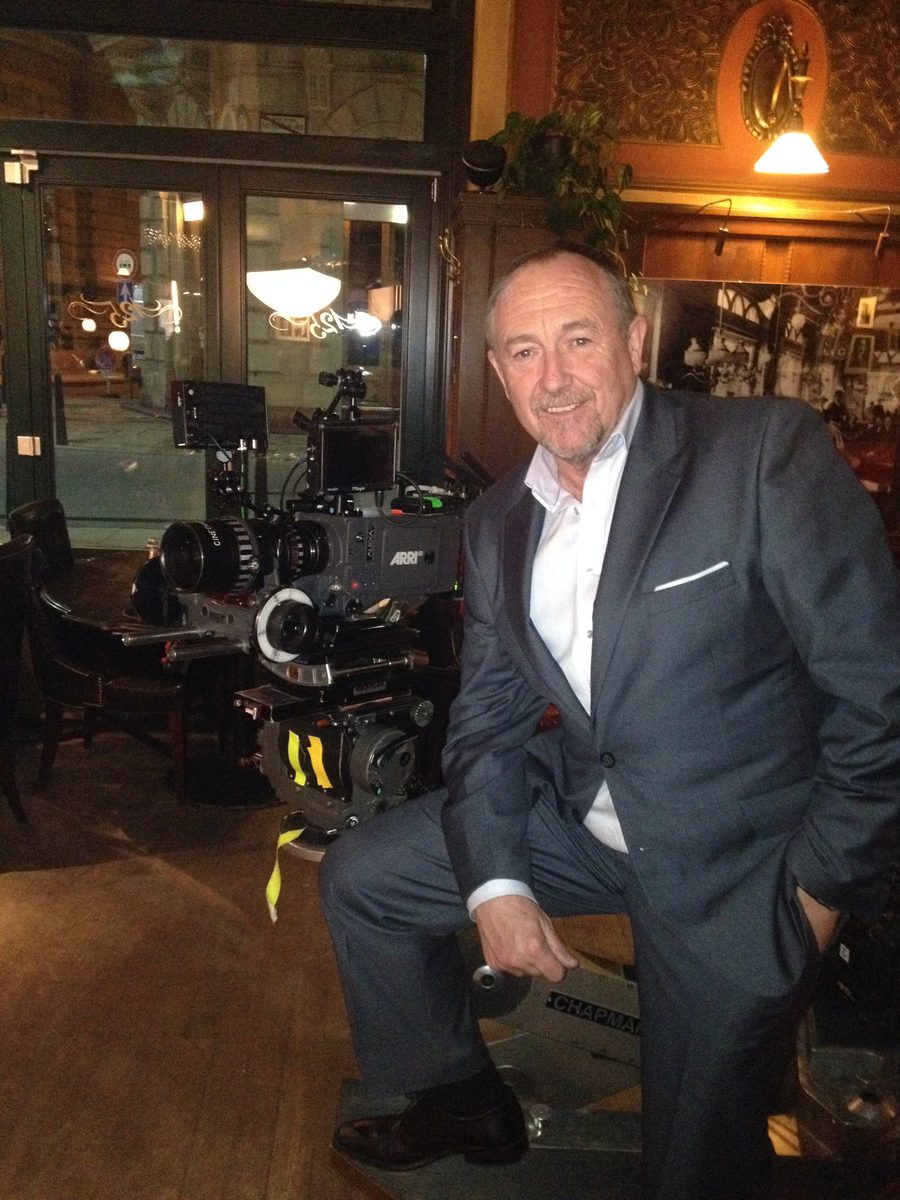 Mark Whelan in a suit beside a film camera on a bar set