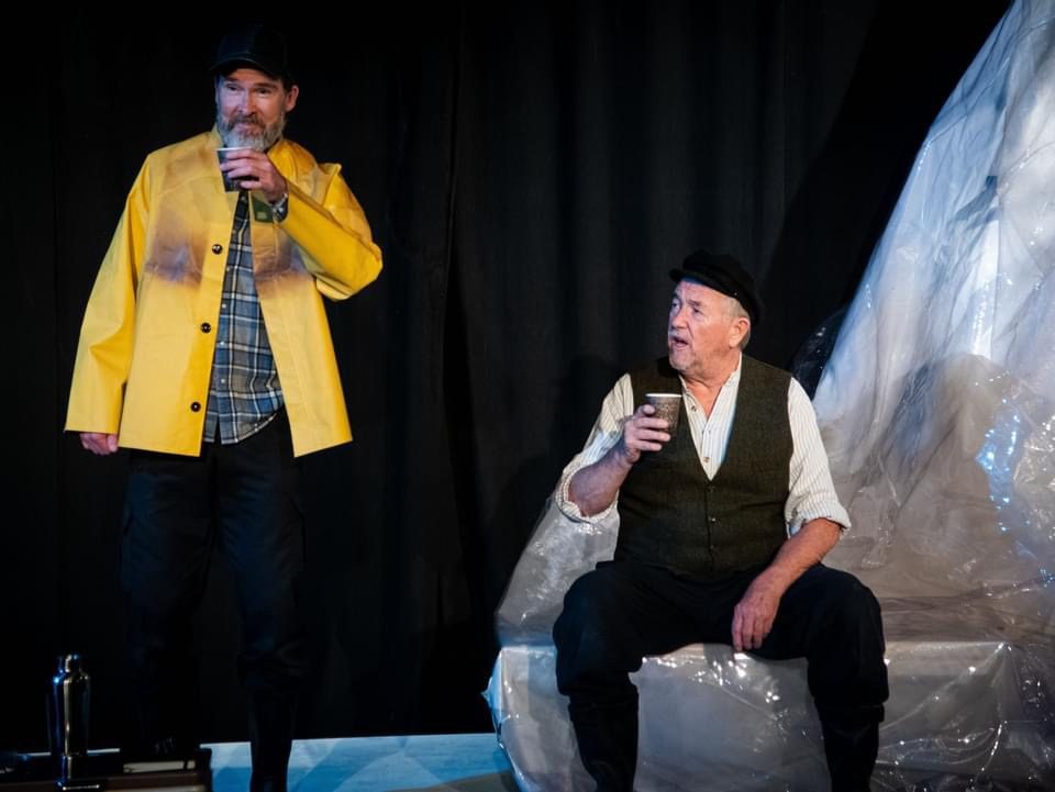 Mark Whelan seated with a cup while an actor in a yellow coat stands beside him